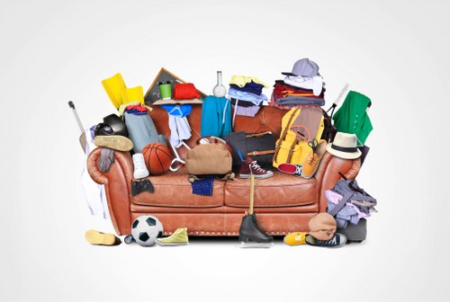 A collection of various discarded household items piled on and around a light brown fabric sofa, including clothing, sports equipment such as a soccer ball and basketball, shoes, a hat, a small cardboard box filled with miscellaneous objects, and a yellow plastic container. Items are arranged haphazardly, with some draped over the sofa arms and others resting on the seat cushions, creating a cluttered scene indicative of accumulated rubbish. The background is plain white, emphasizing the contrast between the objects and the clean backdrop. This image reflects a typical scenario of domestic rubbish accumulation requiring disposal or clearance services, relevant to companies like Office Clearance Hatch End operating within the Hatch End area or nearby postcode regions. The varied textures and colors show the mix of materials common in household waste, from soft textiles and sports gear to hard plastics and cardboard, illustrating the type of refuse handled in a typical rubbish removal process.