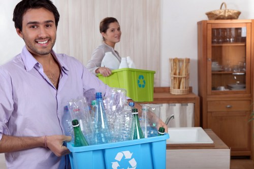A man with dark hair and a beard, wearing a light purple shirt, is holding a blue recycling bin filled with empty, clear plastic bottles, some with labels still attached. Behind him, a woman with brown hair tied back, dressed in a light grey top, is standing next to a green recycling bin with a blue recycling symbol, positioned on a kitchen countertop. The kitchen features light wood cabinetry, a white sink, and a wooden display cabinet with glass doors and shelves, all illuminated by ambient indoor lighting. The scene appears to be in a residential setting, possibly in Hatch End, with a focus on waste sorting and rubbish disposal, highlighting environmentally conscious practices and the importance of rubbish removal services. Office Clearance Hatch End provides professional waste management solutions to support local households and businesses, ensuring clutter and waste are responsibly cleared from homes and workspaces in the area.