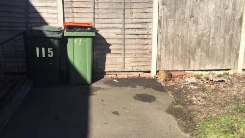 The image shows two large wheelie bins positioned on a paved driveway area, with one bin being dark green and the other a shade of greyish-green, both featuring closed lids. The green bin has the number '115' printed in light-colored font on its side. They are located adjacent to a wooden fence on the right side and a timber panel with horizontal slats behind, suggesting a residential or commercial property boundary. Sunlight casts shadows over part of the bins and the surrounding pavement, which has a damp patch or water stain near the base of the bins. To the right of the bins, the ground transitions from concrete to a small patch of soil with some sparse vegetation, indicating an outdoor environment possibly associated with rubbish or waste collection. The scene portrays an area where rubbish storage occurs, relevant to the services provided by Office Clearance Hatch End for rubbish removal and waste disposal in the local area near Hatch End.