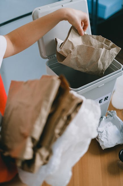 A collection of overflowing rubbish bins and trash on a paved sidewalk in an urban setting, with discarded paper, cardboard boxes, plastic bags, and miscellaneous waste spilling onto the ground. The l