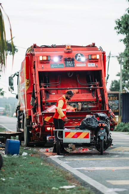 A worker in a high-visibility orange vest is operating a red rubbish collection truck on a roadside, with the back loading area open and debris visible underneath. The worker appears to be managing wa
