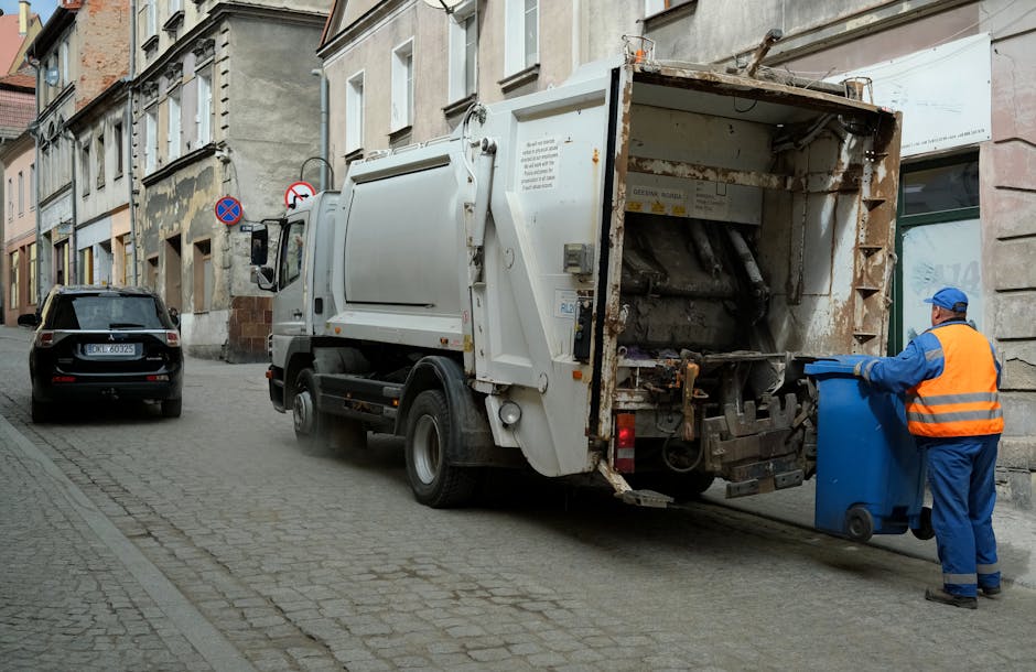 A worker in a high-visibility orange vest is operating a red rubbish collection truck on a roadside, with the back loading area open and debris visible underneath. The worker appears to be managing wa