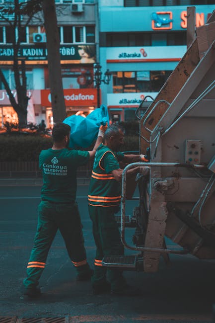 Two waste collection workers wearing high-visibility vests are seen lifting a large blue plastic bag onto a rubbish collection vehicle parked on an urban street. The worker on the left stands upright