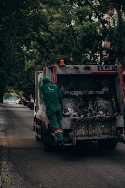 Two waste collection workers wearing high-visibility vests are seen lifting a large blue plastic bag onto a rubbish collection vehicle parked on an urban street. The worker on the left stands upright