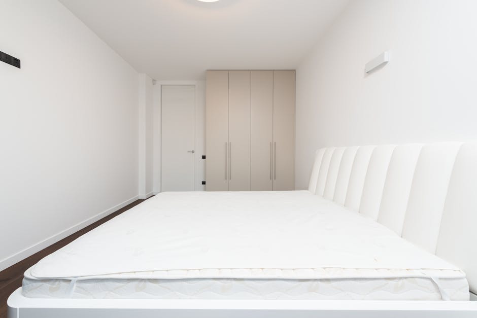 A modern bedroom featuring a neatly made bed with a white mattress, positioned against a textured brick accent wall on the left side. The bed has a grey upholstered headboard, with a small round bedsi