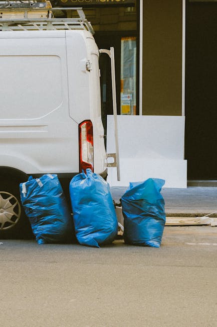 A waste collection operative, dressed in a yellow high-visibility vest with red accents, is seen standing next to the rear of a large red rubbish truck parked on the side of a street. The operator is